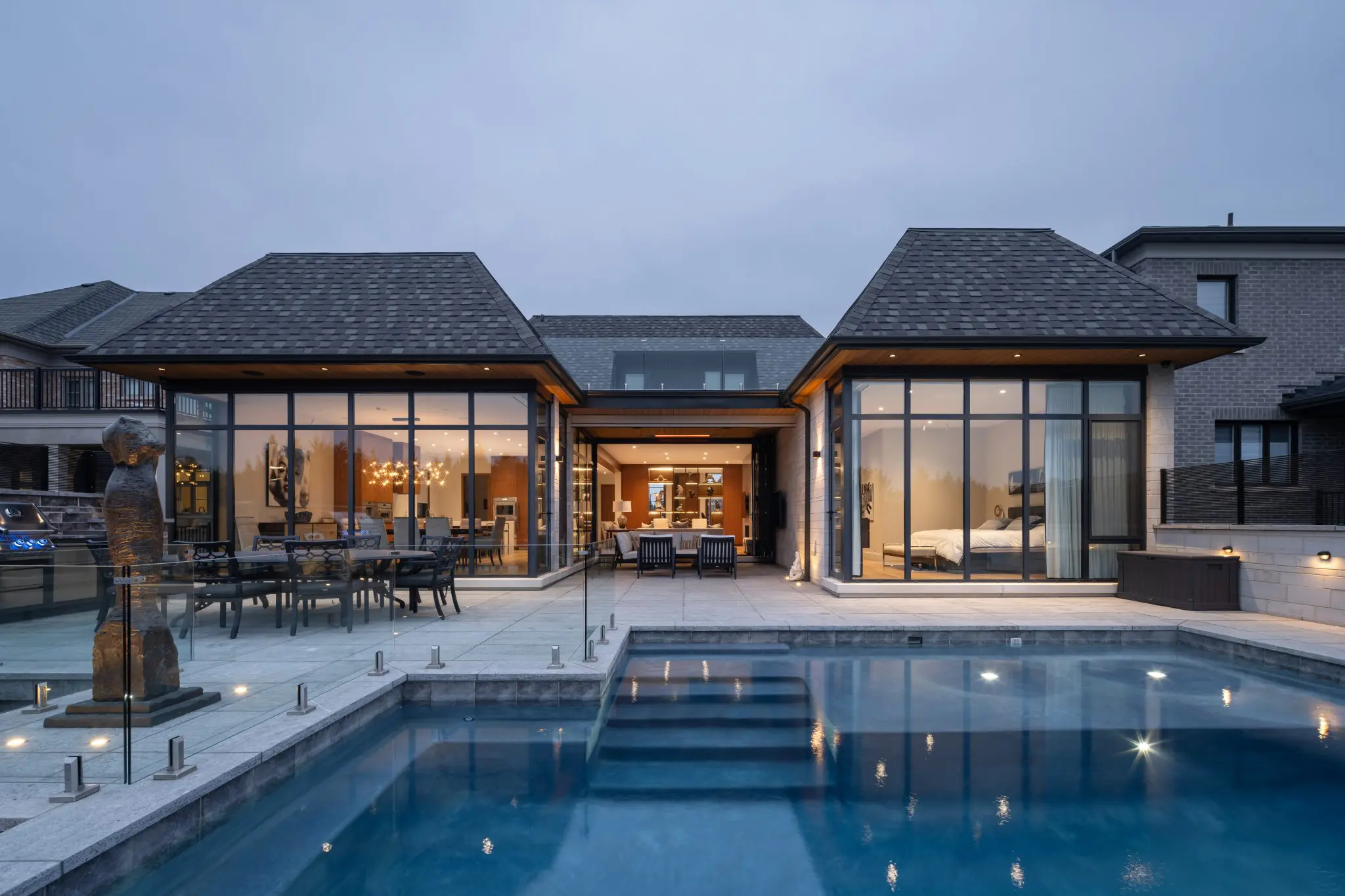 Rear exterior with large glass walls opening to illuminated pool and patio at dusk.