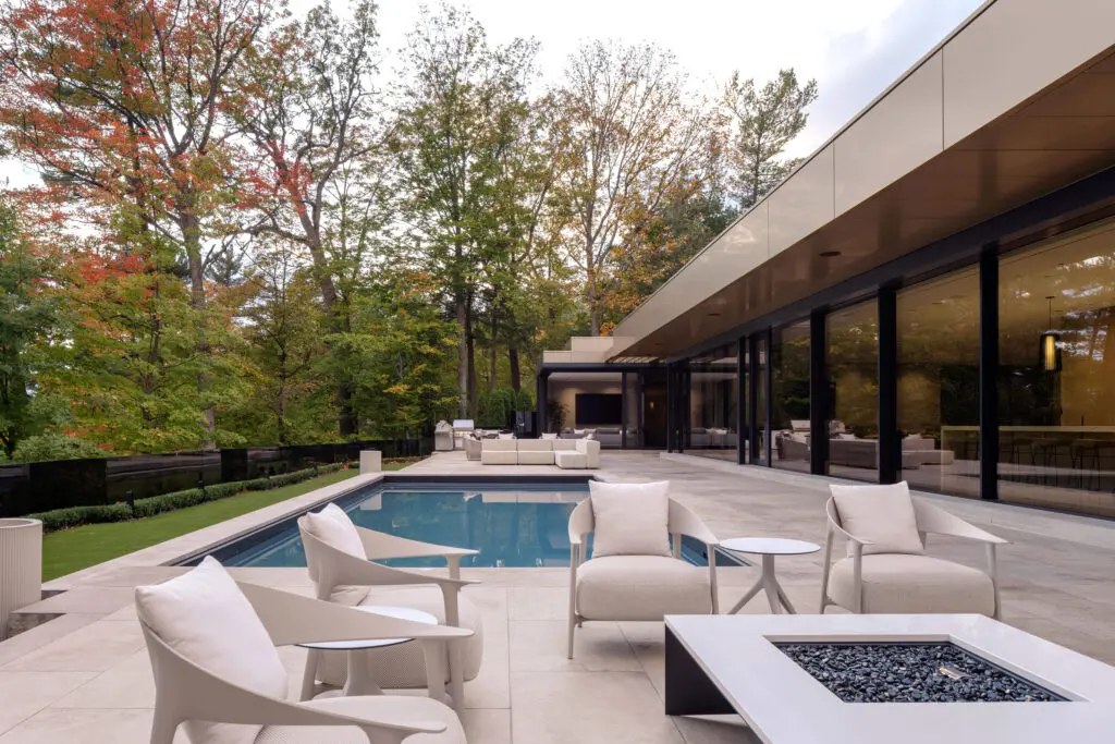 Modern outdoor terrace with pool, lounge seating, and forest backdrop beside glass-walled home.