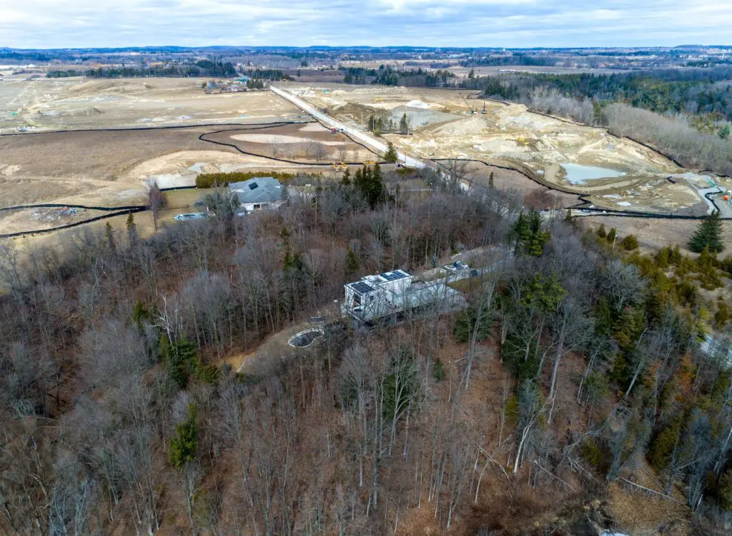 Aerial view of modern home set within wooded landscape and surrounding rural development.