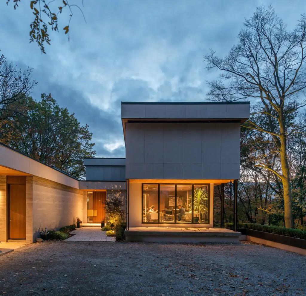 Modern home exterior at dusk with warm interior lighting and glass walls set within wooded landscape.