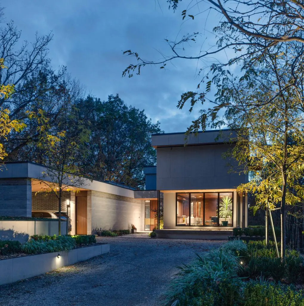 Contemporary residence at night with illuminated entry, landscaped driveway, and forest surroundings.