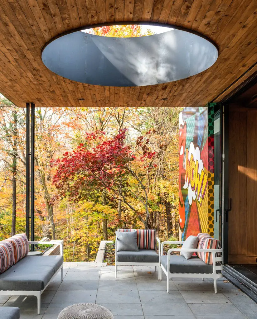Covered outdoor patio with circular skylight, lounge seating, and forest views beside pop art mural.