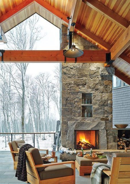 A cozy indoor outdoor living space featuring a Vermont Mountain House with a brick fireplace that opens the room up to the outdoors with tall trees.