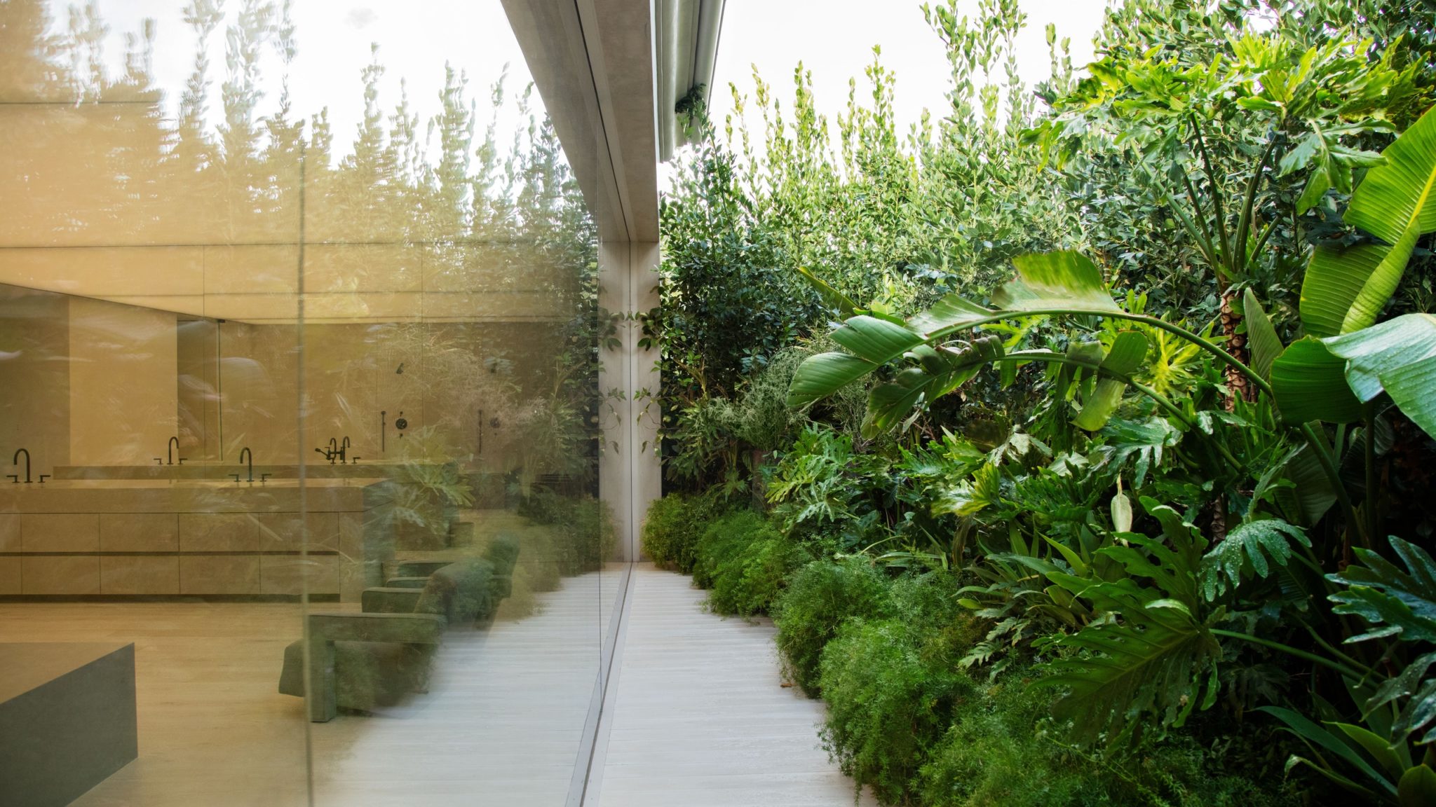 A bright outdoor indoor bathroom with a seamless floor to ceiling glass window and lush greenery surrounding the space. This is the Kardashian-West's newly renovated master bathroom.